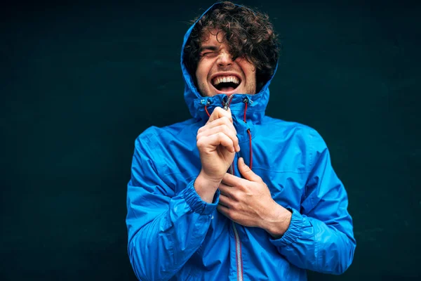 Happy young man smiling broadly, wearing blue raincoat during rain outside. Cheerful male in blue raincoat enjoying the rain on black wall outdoor. Young man has joyful expression in rainy weather.
