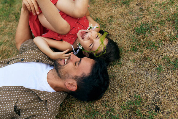 Smiling father playing with his happy daughter during lying on the grass outdoors. Dad enjoying at day out with his child in the park. Daddy and little girl having fun together Happy father's Day. 