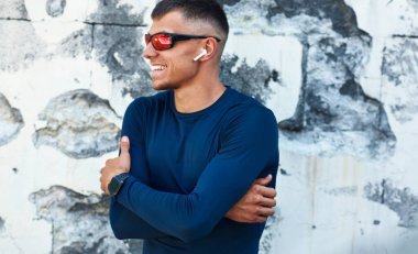 Açık havada mavi spor giyim ve kırmızı güneş gözlüklü gülümseyen bir sporcunun portresi uzağa bakıyor ve beton bir duvarın önünde duruyor. Sporcu kolları dışarıda karşıya geçerken dinleniyor..