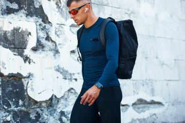 Mavi spor kıyafetli ve güneş gözlüklü bir adamın şehir caddesinde yürüyüşünün yan görüntüsü. Sporcu spor salonuna gidiyor..