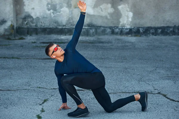 一名男子运动员在城市街道上伸展身体的全景 身穿蓝色运动服 头戴眼镜 戴耳机在城市混凝土墙外锻炼的健身男子 — 图库照片