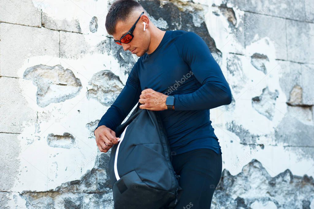 Athletic man in blue sportswear and red sunglasses opening his backpack for a bottle of water standing against concrete wall in the city street. Sportsman going to the gym.