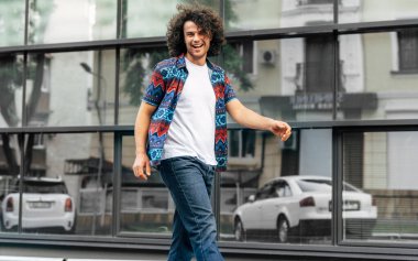 Kıvırcık saçlı neşeli bir adamın şehir caddesinde yürürken yüzünde neşeli bir ifade var. Neşeli, güler yüzlü, modaya uygun kıyafetler giyen erkek. Kasabanın güneşli yaz gününün tadını çıkarıyor..