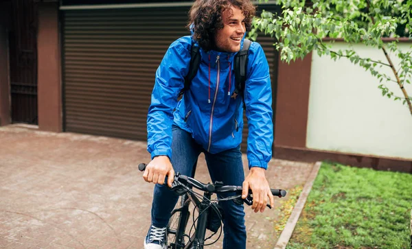 Handsome curly man cycling on his bike down the street next to the house. Cheerful male courier in blue rain coat with curly hair delivers parcel cycling with a bicycle in the city in a rainy day