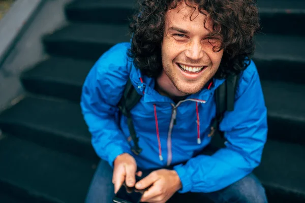 Mavi Yağmurluklu Gülümseyen Genç Adamın Portresi Merdivenlerde Oturan Cep Telefonunu — Stok fotoğraf
