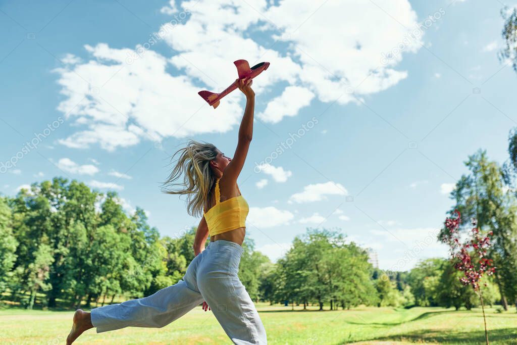 Beautiful young woman running in the park while playing with toy airplane on a sunny day outdoor. Side view female jumping and holding an airplane ready to set it free to fly. Travel concept.