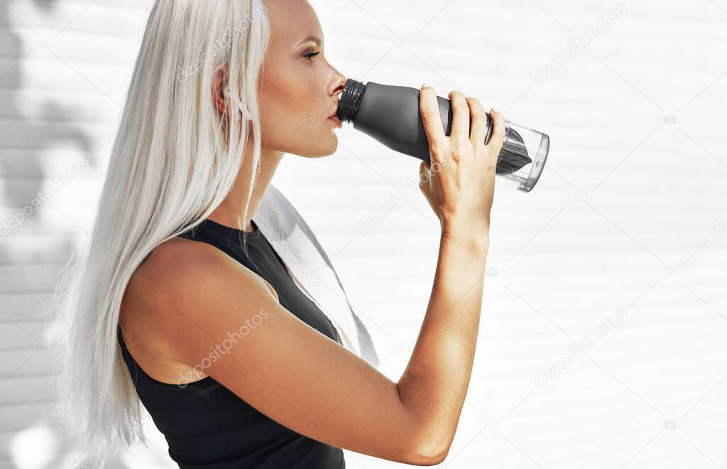 A beautiful blonde female runner standing against white wall drinking water from the bottle. Fitness girl taking a break after the training session drinks the detox water.