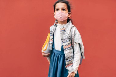 Korunaklı yüz maskesi takan güzel bir çocuğun koronavirüs sırasında okula dönüşünün yatay dış görüntüsü. Sırt çantası ve klasörüyle dışarıda poz veren sevimli liitel kız öğrenci..