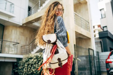 Sırt çantalı ve kitaplı rahat giyimli, gözlüklü güzel bir kadının arka plan görüntüsü. Üniversite kampüsünde kitap taşıyan uzun kıvırcık saçlı güzel hippi kız öğrenci..