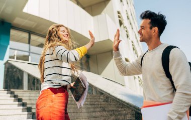 Dışarıdaki üniversitenin yanında beşlik çakan iki genç öğrencinin yan görünüşü. İki meslektaş, genç bir kadın ve genç bir adam açık hava kampüsünde buluştular..