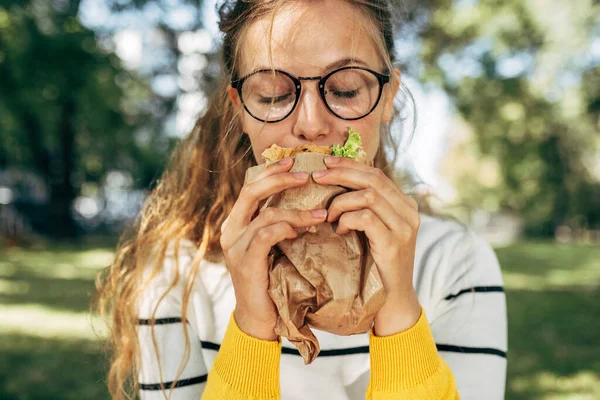 学生の女性のクローズアップ肖像画は 屋外でサンドイッチと昼食を持っています 眼鏡のブロンドの若い女性は公園でファーストフードを食べるために休息を取る — ストック写真