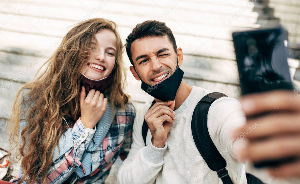 Image of two happy young students taking selfies on the mobile phone sitting on the stairs of the college campus. Smiling woman and man in protective face masks online conversations on the smartphone.