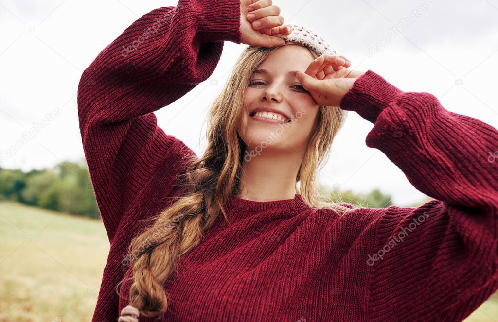 Happy young blonde woman in red sweater and winter hat smiling broadly posing on nature and sky background. 