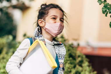 Koronavirüs sırasında okula giderken pembe koruyucu maske takan mutlu bir çocuk. Sırt çantalı, gülümseyen küçük bir kız öğrenci sokakta yürüyor..