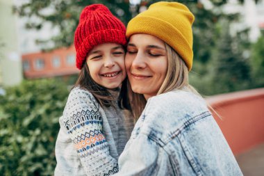 Kırmızı şapkalı mutlu küçük kızın portresi sarı şapkalı, kot ceketli annesine sarılıyor. Annesini kucaklayan sevimli çocuk dışarıda eğleniyor. Anne ve kızı sevgiyi paylaşıyor. Anneler Günü.