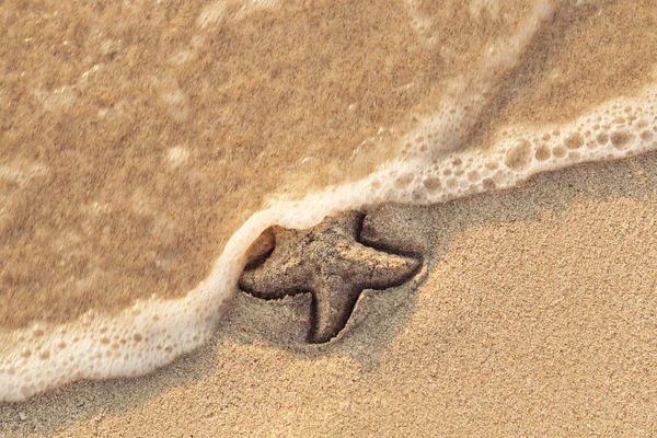Estrellas de mar dibujadas en la arena de la playa siendo arrastradas por una ola. Ola de mar espumosa viene a lavar una imagen en arena de playa de color amarillo húmedo. Mensaje festivo y vocacional. Vista desde arriba. Copiar espacio . — Foto de Stock