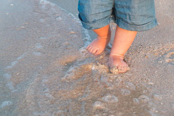 Bare feet walking at sandy beach near the sea. Little baby in blue jeans shorts going to touch the sea at sunset. Wave washes baby's feet. Toned. Soft focus