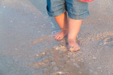 Çıplak ayak, kumsal deniz kenarında yürüyüş. Küçük bebek kot şort gün batımında denize dokunacak. Dalga bebeğin ayakları yıkar. Tonda. Yumuşak odak
