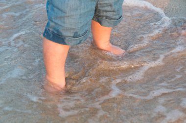 Çıplak ayak, kumsal deniz kenarında yürüyüş. Küçük bebek kot şort gün batımında denize dokunacak. Dalga bebeğin ayakları yıkar. Tonda. Yumuşak odak