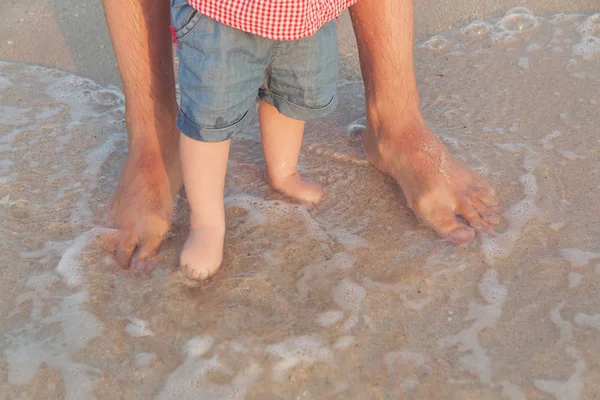 Dalga için bekleyen sığ suda duran adam ve bebek ayaklar. Çıplak ayak babası ve onun küçük kızı veya oğlu kum denize yakın kalmak. Seyahat ve tatil kavramı. Tonda. Yumuşak odak
