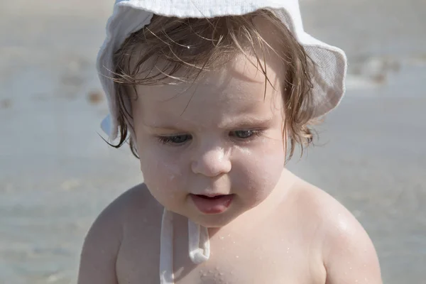 Cute sad baby with sunscreen cream on cheek against sea background. Pretty infant girl in white hat and with sunscreen and water drops on her face playing at the beach. Safety in summer sun