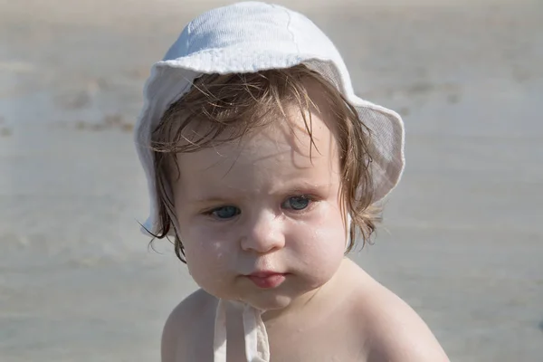 Cute sad baby with sunscreen cream on cheek against sea background. Pretty infant girl in white hat and with sunscreen and water drops on her face playing at the beach. Safety in summer sun