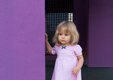 Pembe yaz aylarında şirin bebek elbise bezelye 1 yaşında açık havada poz içinde. Uzağa arıyorsunuz. Çocukluk. Ciddi ve güzel küçük sarışın kız mavi gözleri modeli ayakta mor duvarının yakınında iyi giyimli portresi. Sevimli çocuk bebek giysileri içinde poz