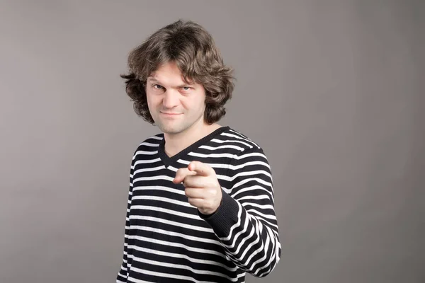 Joven con suéter a rayas con el pelo peludo, el dedo apuntando directamente hacia la cámara con el dedo índice, te culpa de hacer cosas malas sobre un fondo gris. Oye, tú. Captura de estudio — Foto de Stock