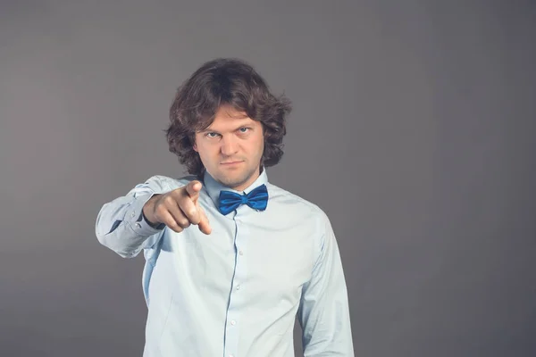 Young man in blue shirt with bow tie with shaggy hair, finger pointing directly towards the camera with index finger, blames you in doing wrong things on a gray background. Hey you. Studio shot.