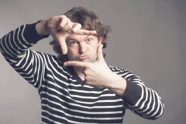 İle çerçeveleme parmakları ile odaklanarak genç yakışıklı shaggy kıvırcık adam gri arka plan üzerinde kopya alanı eller. Portre adam Çizgili kazaklı bir çerçeveleme hareketle ellerini uzatarak kapatın