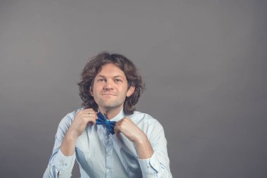 Onun papyon içinde ayarlama genç erkek hipster stüdyo çekim üzerinde izole gri arka plan ayna. Siyah kıvırcık saçlı damat gelin ile Toplantı için hazırlanıyor mavi gömlekli. Kameraya bakarak ve gülümseyen.