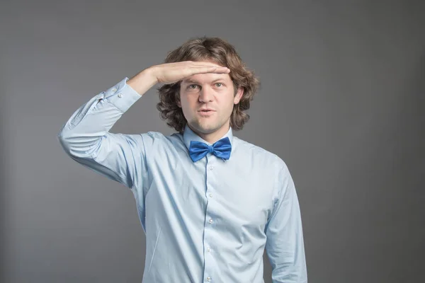Handsome young man with dark curly hair wearing blue shirt and bow tie putting hand on his forehead and smiling, looking far isolated on grey background. Looking or waiting for someone or something.
