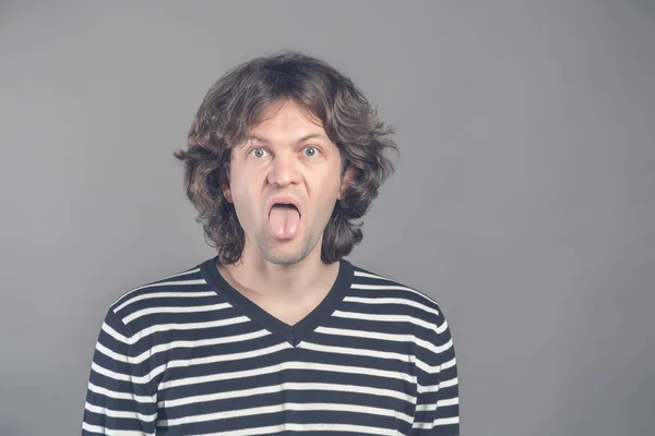 Primer plano retrato de divertido shaggy loco guapo hombre en suéter a rayas mirando a la cámara con la gran boca abierta y la lengua hacia fuera, se burla. Amplio abierto ojos estudio interior disparo, aislado sobre fondo gris . — Foto de Stock