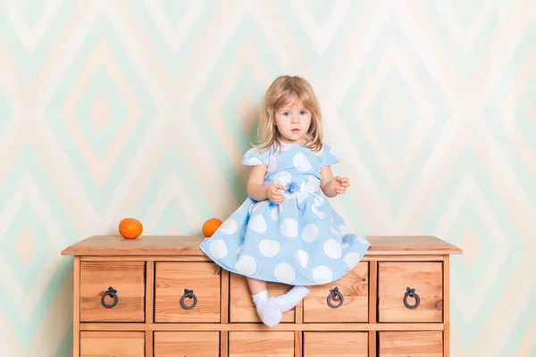 A little baby girl in her room sitting cross-legged on chest of drawers with tangerines on the rhomb wallpaper background. Child in blue polka dot dress and white socks attentively looking at camera.