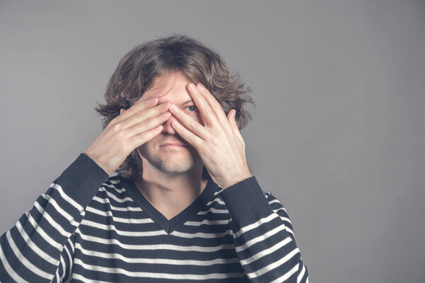 Portrait of young hipster man in sweater peeping at the camera through his fingers. Picture of embarrassed male looking through his hands covering his face isolated against studio interior background.