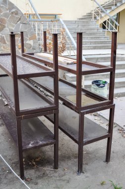 Metal raflar dışarıya, bazı basamakların yanına ve bir duvara yerleştiriliyor. Rafların kahverengi bir rengi var ve muhtemelen depolama veya görüntüleme için kullanılıyor..