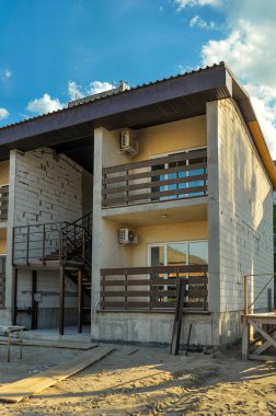 A two-story building under construction with balconies and a dark roof against a bright blue, cloudy sky. The construction area is visible in the foreground.