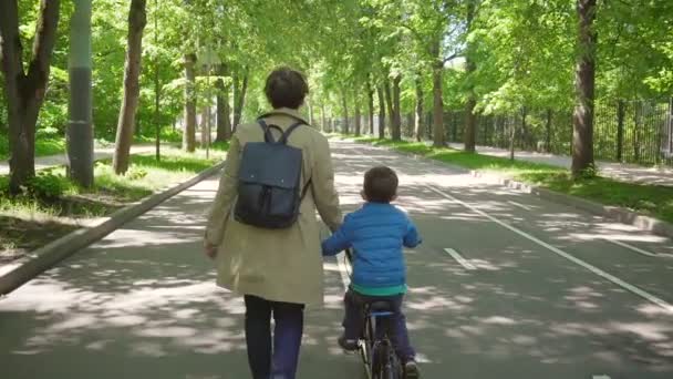 Маленький хлопчик на велосипеді з мамою, що йде в міському парку — стокове відео