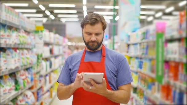 Penjual tampan menggunakan tablet digital berdiri di antara rak-rak di supermarket — Stok Video