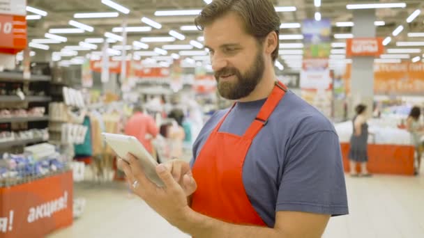 Pěkný supermarket úředník pomocí dotykové obrazovky tabletu v supermarketu, on se usmívá na kameru — Stock video