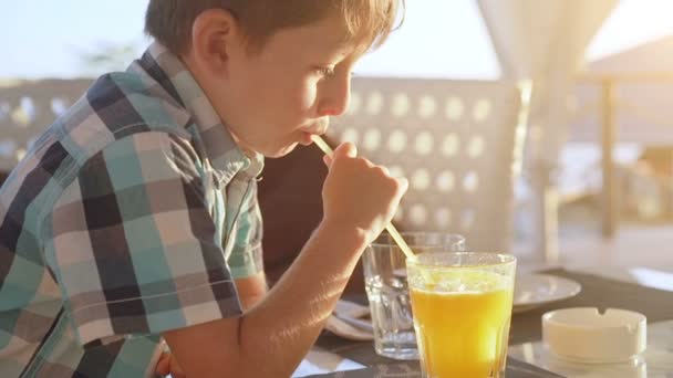 Lindo niño bebiendo jugo de naranja fresco de vidrio en la cafetería de la ciudad — Vídeo de stock