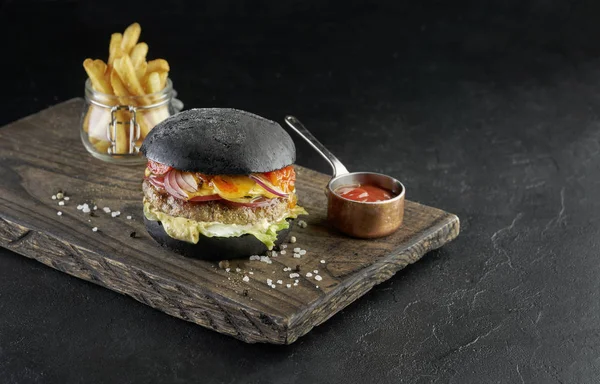 Schwarzer Burger auf rustikalem Holzbrett mit Pommes auf schwarzem Hintergrund. — Stockfoto