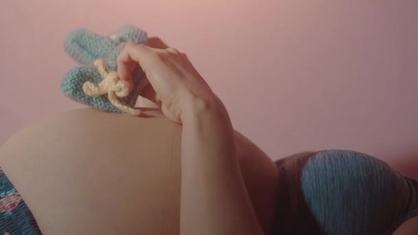 Vientre de una mujer embarazada, las manos de la madre sosteniendo zapatos azules del bebé — Vídeo de stock