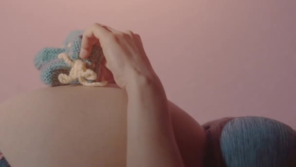 Schwangere liegt auf dem Rücken und tritt mit blauen Babyschuhen auf ihren nackten Schwangerschaftsbauch — Stockvideo
