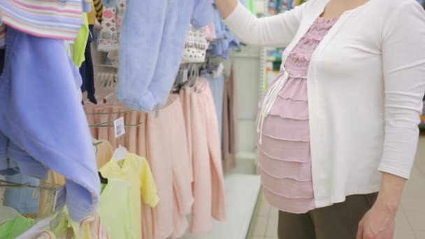 Концепція вагітності - вагітна жінка вибирає блакитний костюм для немовлят в магазині одягу — стокове відео