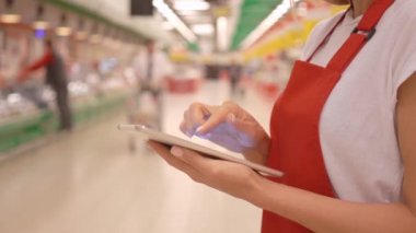 Arka planda müşteriler ve raflar ile dijital tablet kullanarak kırmızı önlük giyen kadın satış memuru
