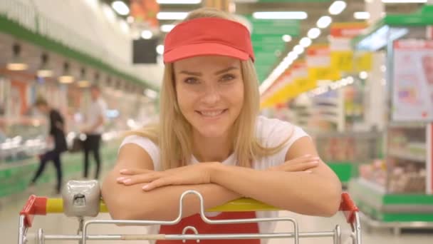 Smiling happy saleswoman dalam seragam merah bersandar pada keranjang belanja di supermarket — Stok Video