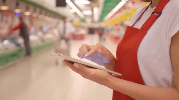 Mujer empleada de ventas con delantal rojo usando una tableta digital con clientes y estantes en el fondo — Vídeo de stock