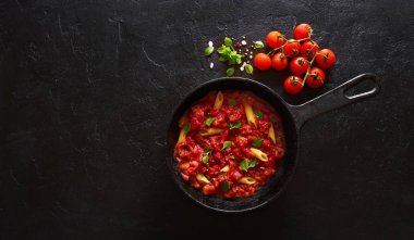 Domates sosu ve yeşil fesleğen siyah bir arka plan üzerinde dökme demir tavada süslenmiş penne makarna. Kopyalama alanı ile üstte görünüm
