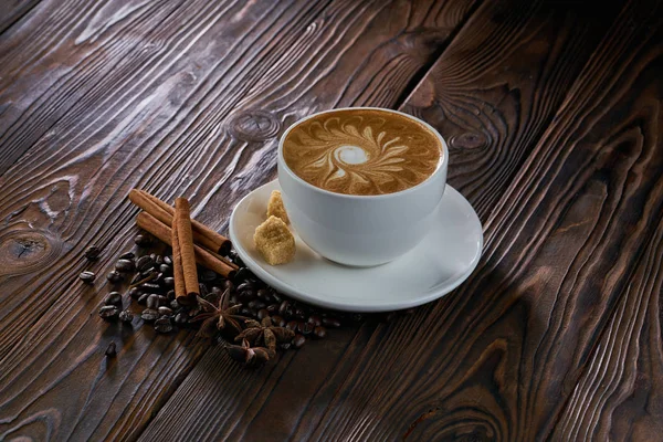 A cup of cappuccino with coffee beans, cinnamon and brown sugar on wooden table — Stock Photo, Image